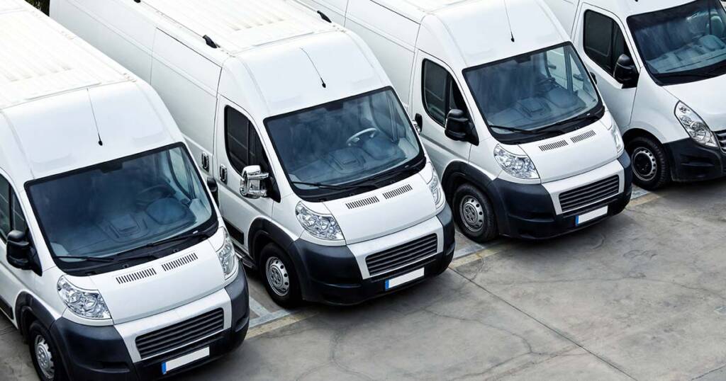 A fleet of identical white cargo vans parked diagonally in a pattern, ready for dispatch. The vehicles are arranged neatly in an outdoor parking lot, highlighting their uniformity and readiness for transportation or delivery services.