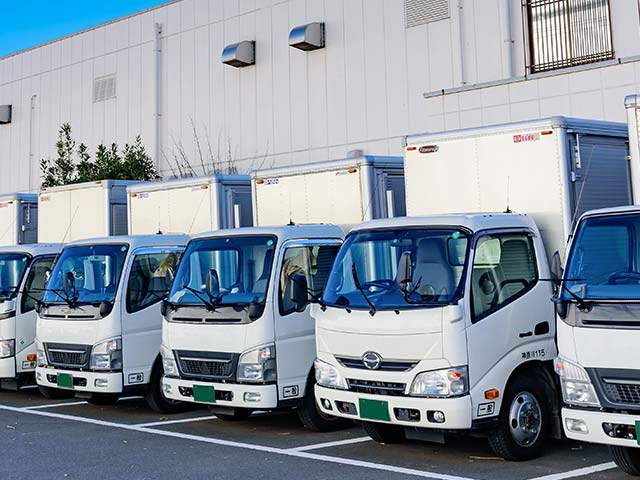 A row of white commercial trucks with cabs in various states of tilt parked in front of a warehouse. Each truck has a distinct front grille design, indicating different makes or models, yet they share a cohesive look for an organized fleet.