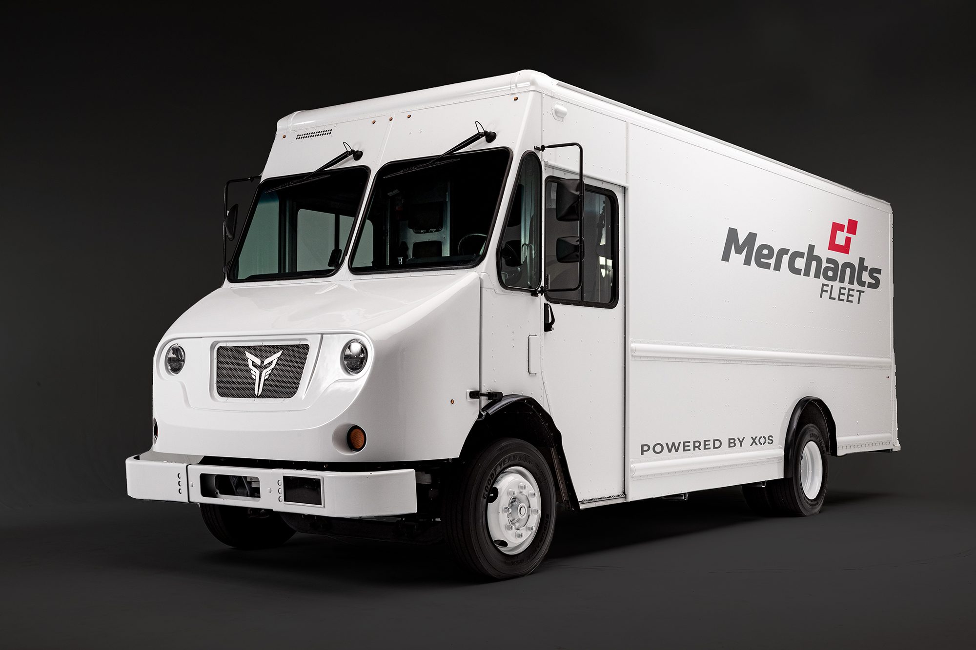 White electric delivery truck with "Merchants Fleet" and "Powered by XOS" logos on a dark background.