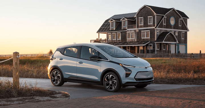 A silver car parked on a driveway near a large, rustic house with a sunset in the background.