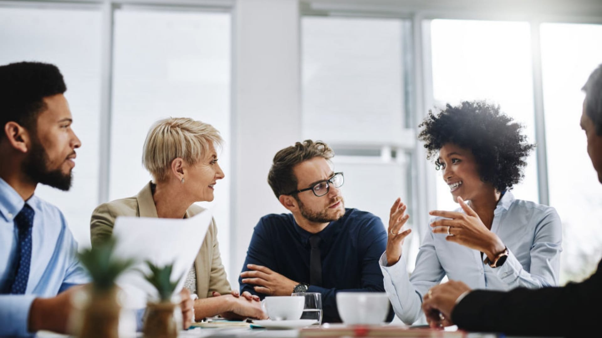 A group of people in a meeting room having a discussion, with papers and coffee cups on the table.