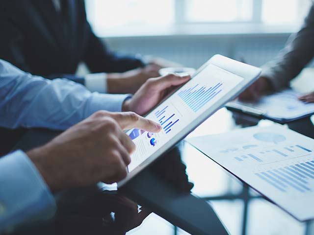 Close-up of hands using a tablet displaying charts during a meeting with papers spread on a table.