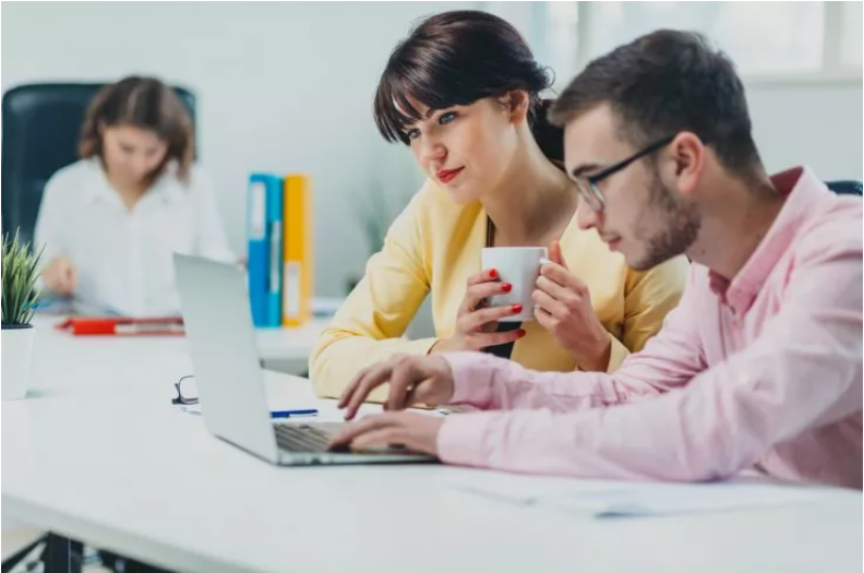 A woman holding a mug and a man working on a laptop, with another person in the background in an office setting.