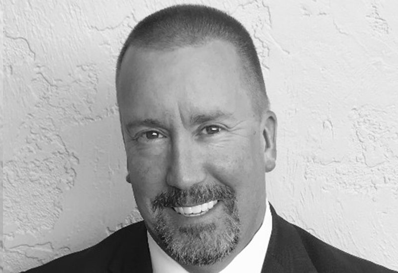 Man with a goatee and short hair, smiling in a suit against a textured white wall.