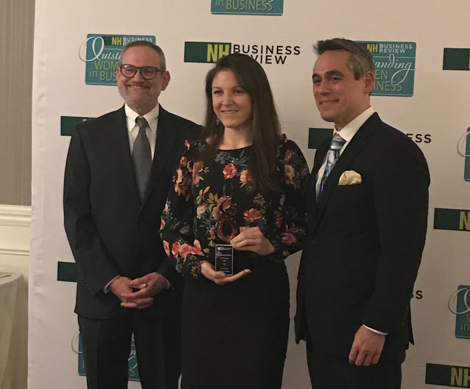 Three people in formal attire posing at an awards event. The woman in the center holds a trophy.