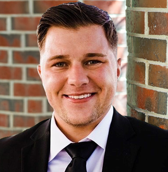 Person in a suit smiling, standing against a brick wall background.
