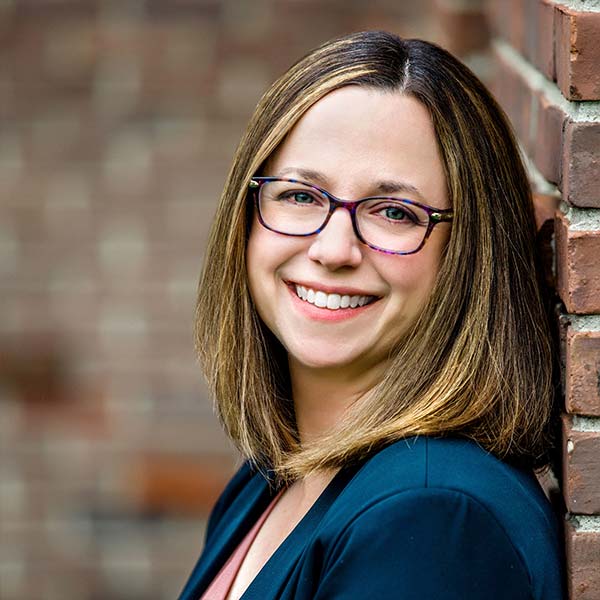Smiling woman with glasses and shoulder-length hair, leaning against a brick wall.
