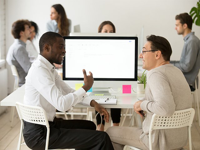 Two colleagues sit and discuss at a desk in an office, with a computer monitor and others working in the background.