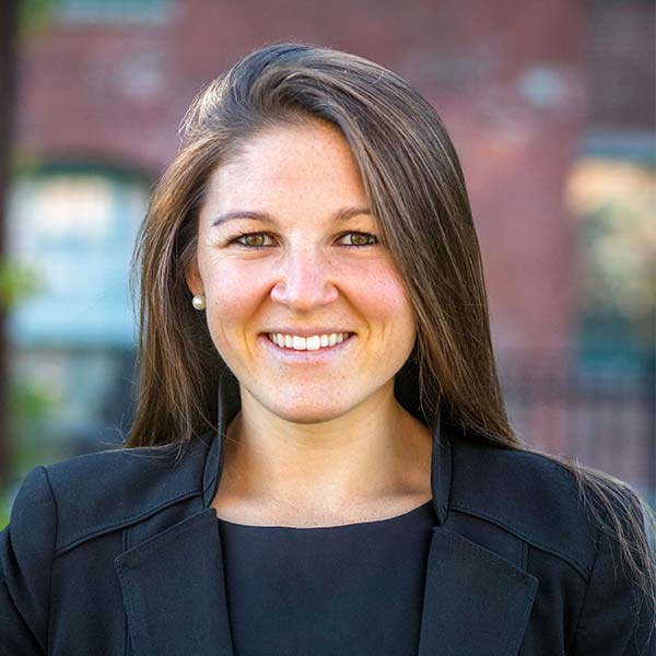 Smiling woman with long brown hair wearing a black blazer, standing outside with a blurred brick background.