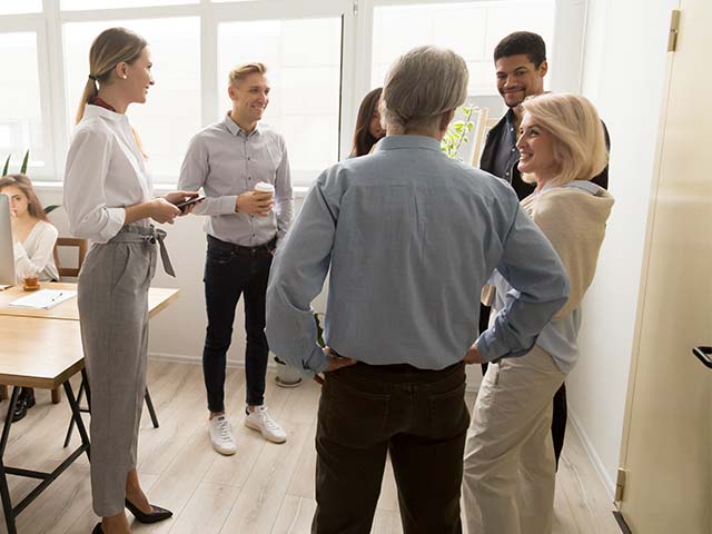 Group of people standing and talking in an office setting, with a woman seated at a table in the background.