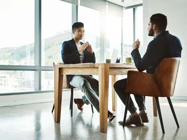 Two people in business attire having a meeting at a wooden table in a bright office with large windows.