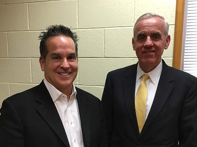 Two men in suits smiling in an indoor setting with a light-colored brick wall background.