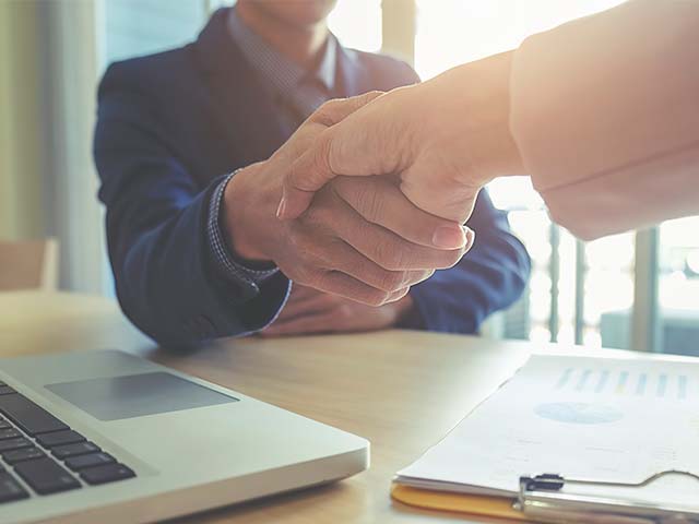 Two people shaking hands over a table, with a laptop and documents visible.