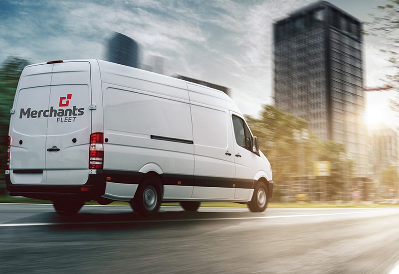White delivery van with "Merchants Fleet" logo, driving fast on city street with blurred buildings in the background.