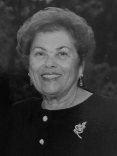 Smiling older woman with short hair, wearing a dark top with a brooch, against a dark blurred background.