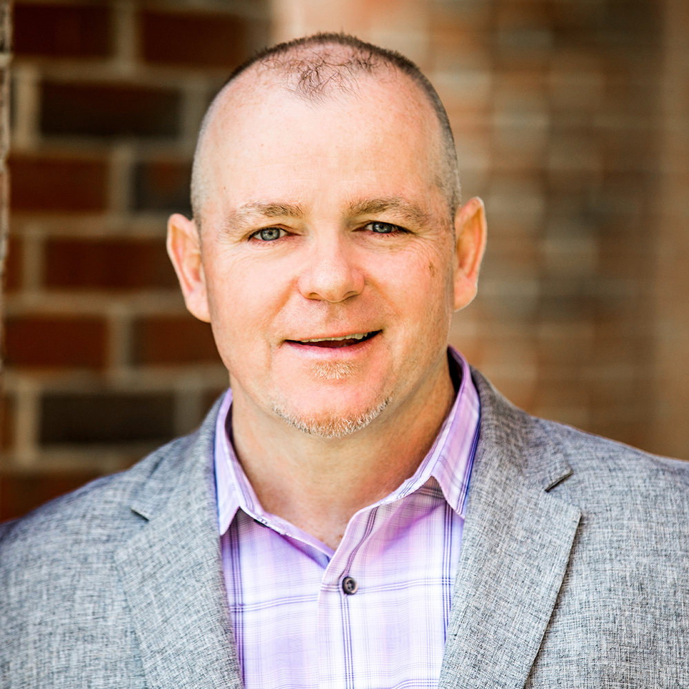 Smiling man in a gray blazer and purple shirt stands against a brick wall.