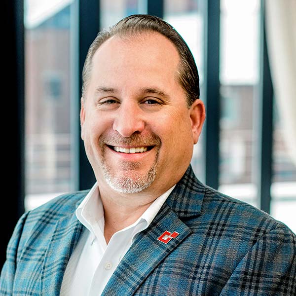 Smiling man in a plaid blazer stands in front of a window with vertical blinds.