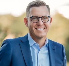 Smiling man in glasses and a blue suit standing outdoors with a blurred background.