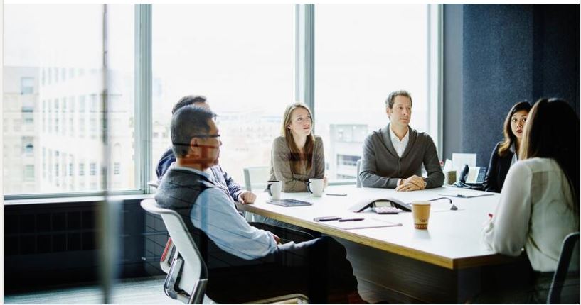 People in a conference room sitting around a table with laptops and coffee cups in front of large windows.