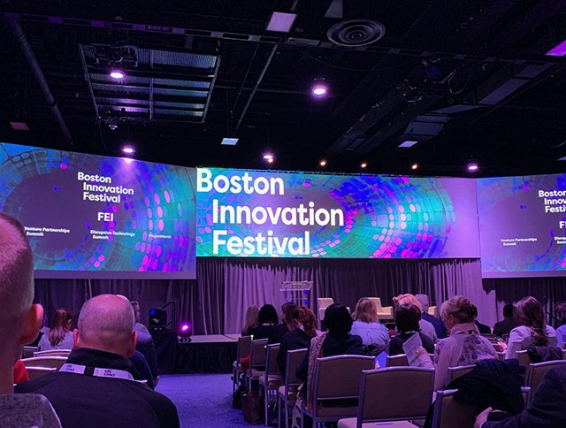 Audience at the Boston Innovation Festival in a dimly lit conference room, view of stage and screens.