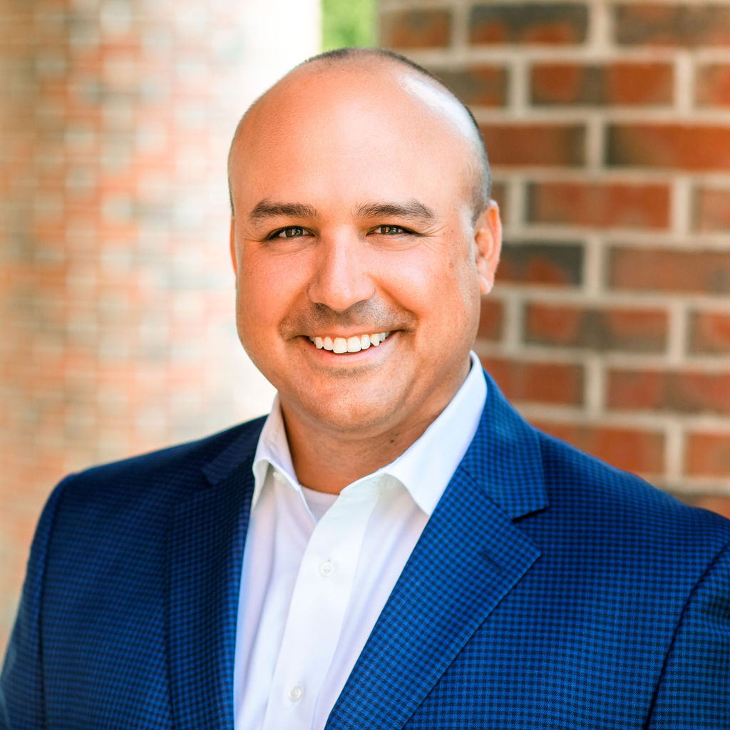 Smiling man in a blue suit and white shirt stands in front of a brick pillar.
