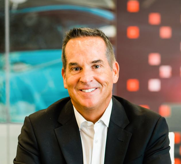 Man in a suit smiling, seated against a background with red and black square patterns.