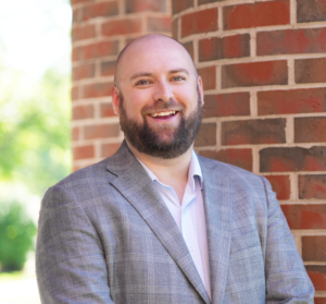 Smiling man with a beard in a gray suit stands next to a brick wall, with greenery in the background.