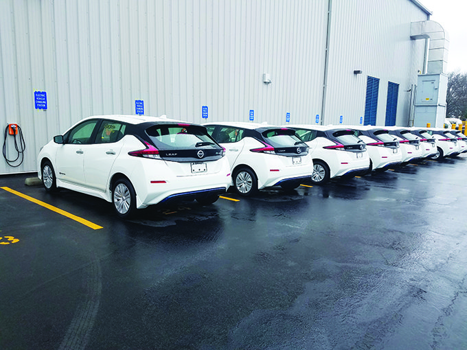 Several white electric cars parked in a row beside a gray building on a wet pavement.
