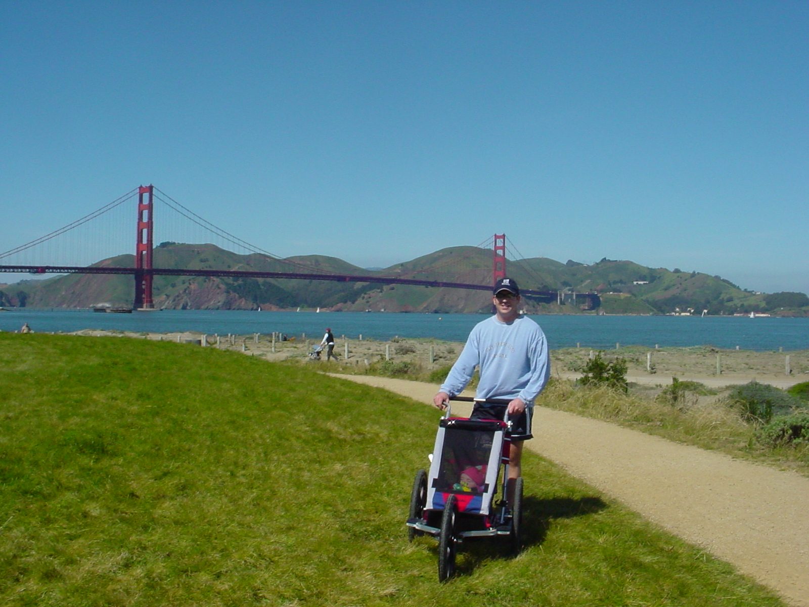 Person with a stroller on a path near a body of water, with a bridge and hills in the background.