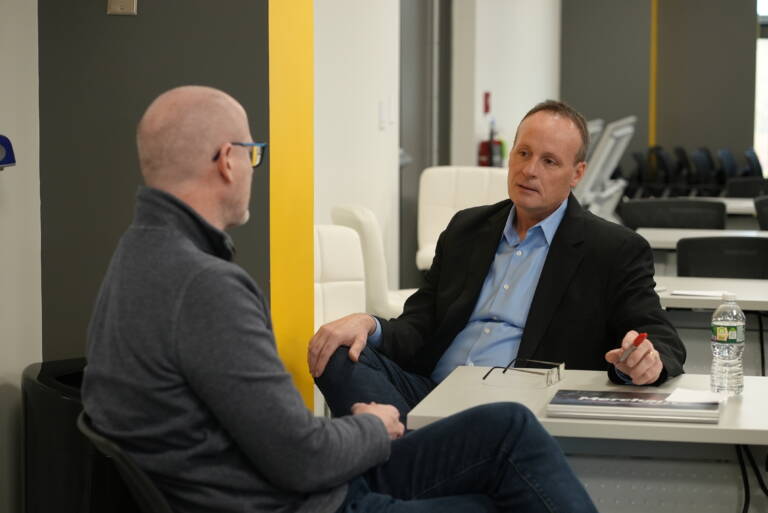 Two men in conversation at a table in an office setting; one holds a pen, the other listens attentively.