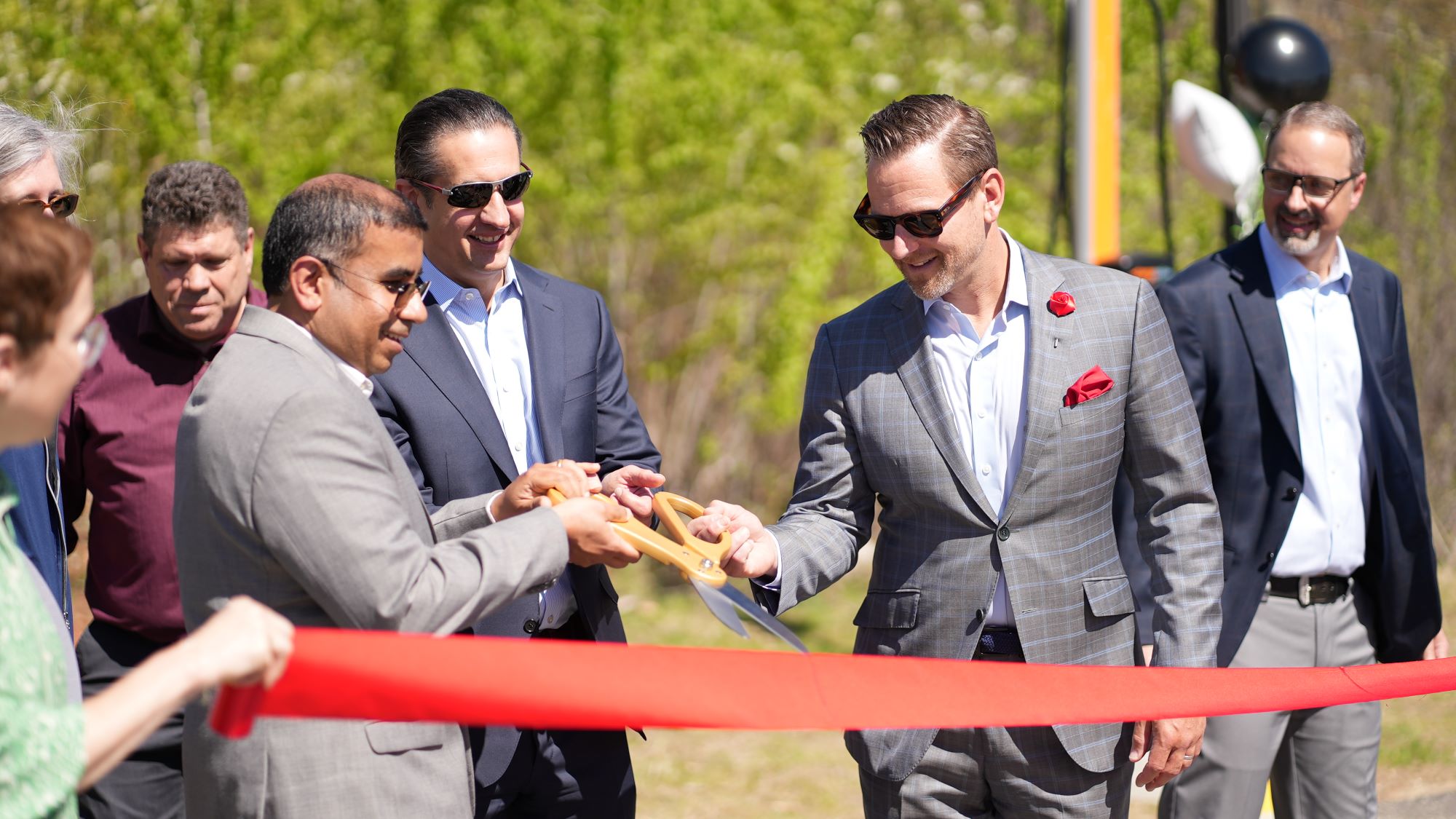 Group of people in suits cutting a red ribbon outdoors with large scissors.