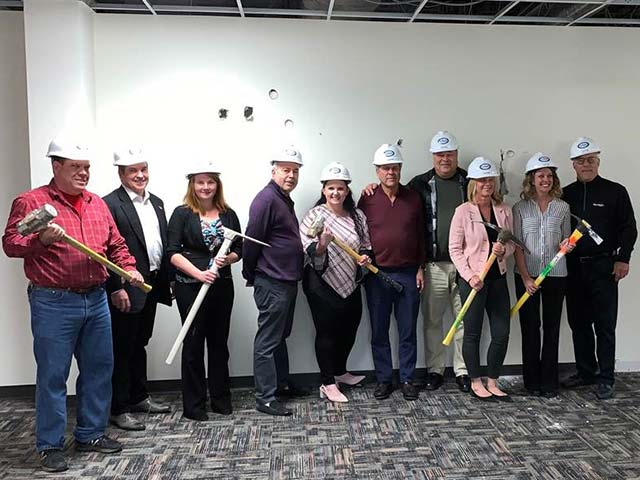 Group of people wearing hard hats, holding sledgehammers, posing in a partially constructed room.