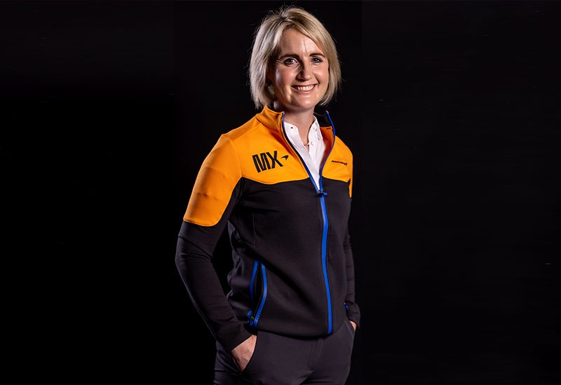 Person in an orange and black jacket with blue accents, smiling against a black background.