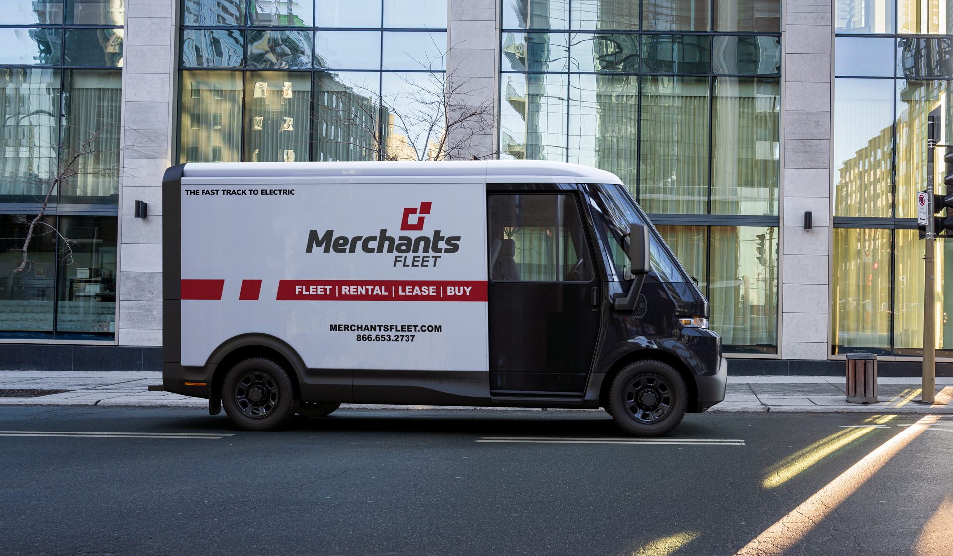 Electric delivery van with "Merchants Fleet" branding parked on a city street in front of a modern building.