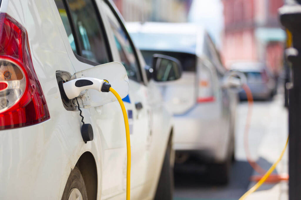 Two electric cars charging on a street, plugged into charging stations with yellow cables.