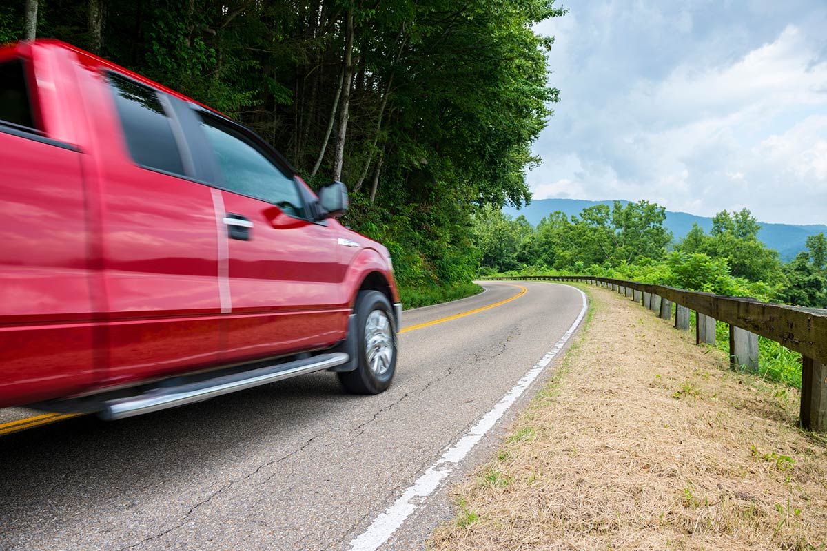 Red pickup truck driving on a winding road through a lush green landscape with trees and cloudy sky.