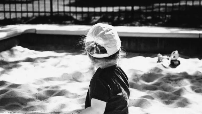 Child in a cap playing in a sandbox with a fence in the background, in black and white.