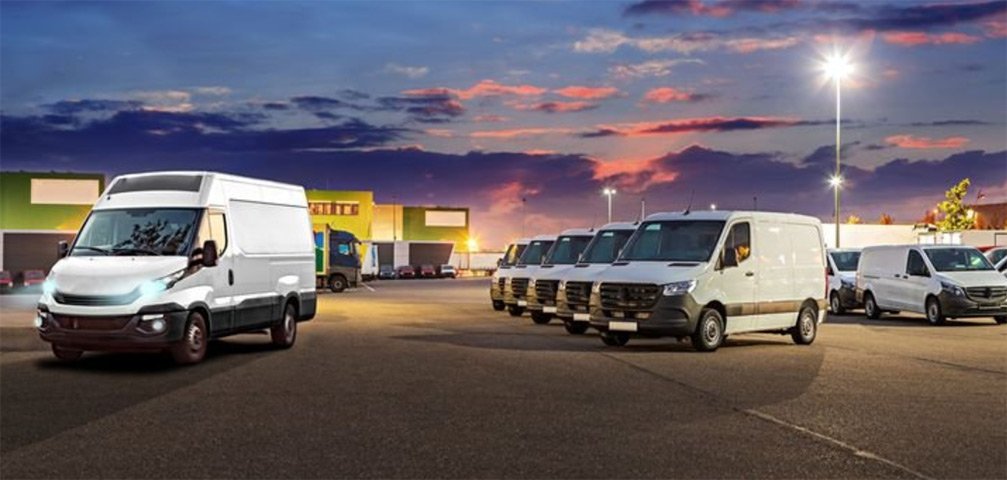 Several white vans parked in a lot at sunset, with one van driving past a row of parked vans.
