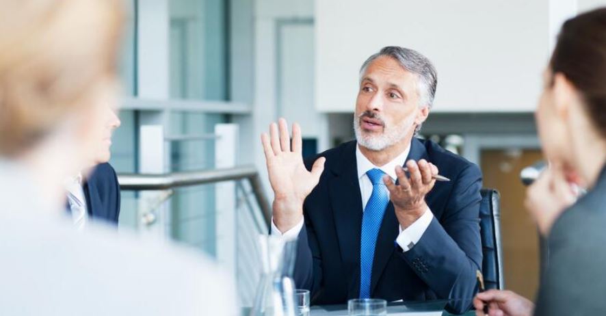 A man in a suit gestures while speaking to colleagues around a table in a modern office setting.