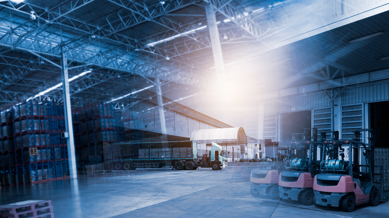 Interior of a large warehouse with stacked pallets, forklifts, and a truck near a loading dock under bright lights.