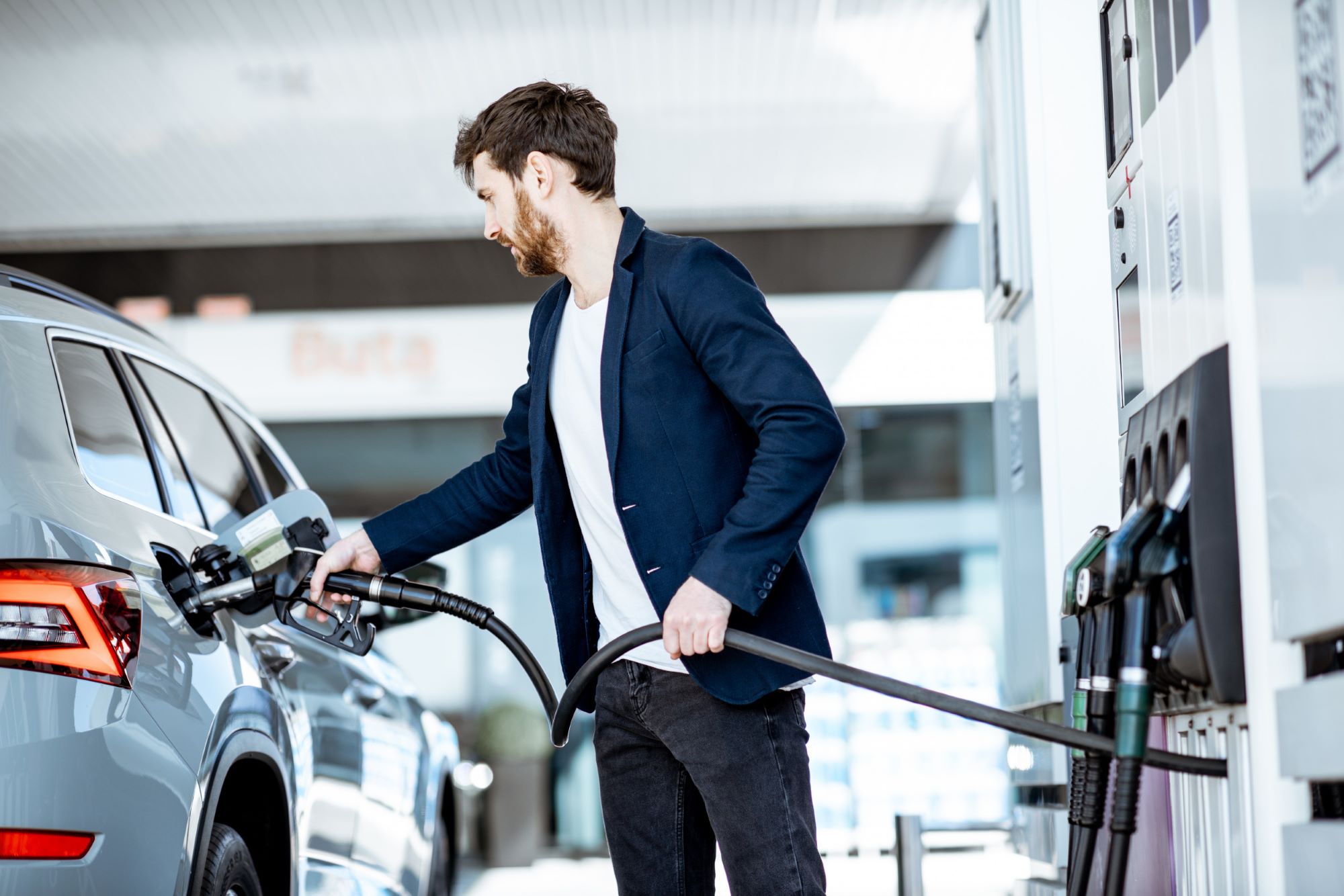 Man refueling a car at a gas station, holding a fuel nozzle in one hand, wearing a dark jacket.