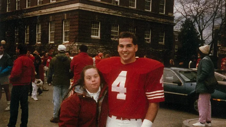 Person in a red football jersey, number 4, standing with a smiling woman in red jacket outside a brick building.