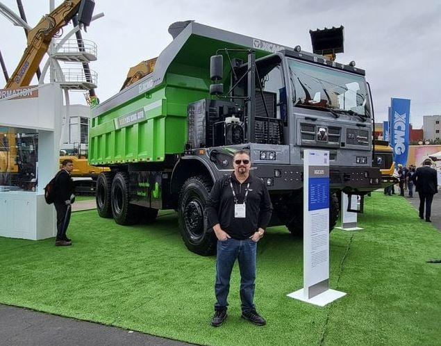 Man standing in front of a large green dump truck at an outdoor exhibition, with people and machinery in the background.