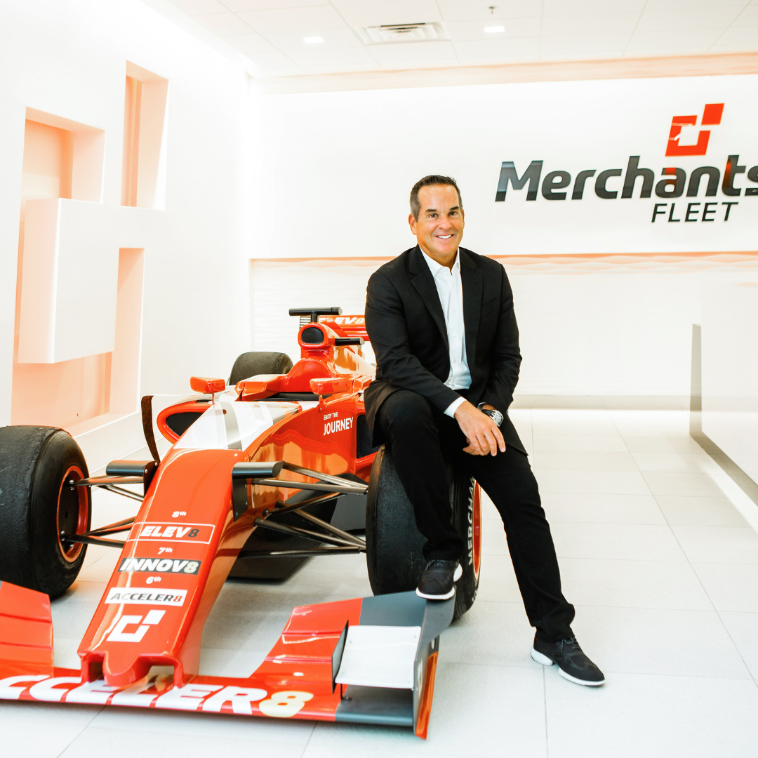 Man in a suit sitting on the tire of a red racing car, with "Merchants Fleet" logo on the wall behind.