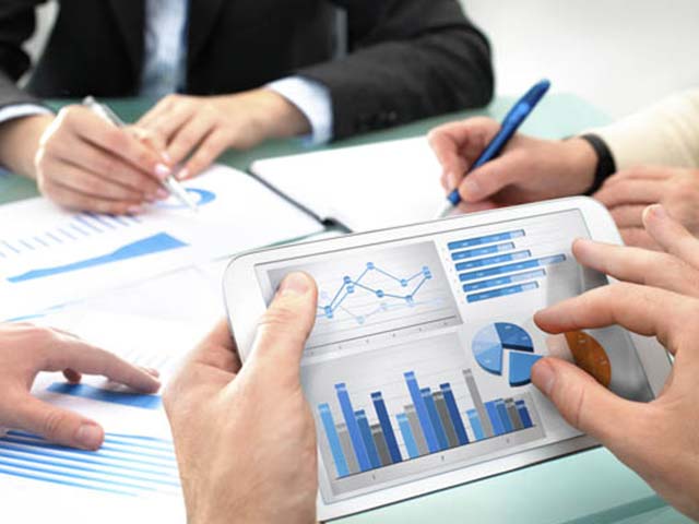 Hands holding a tablet displaying charts, with people writing notes on documents in the background.