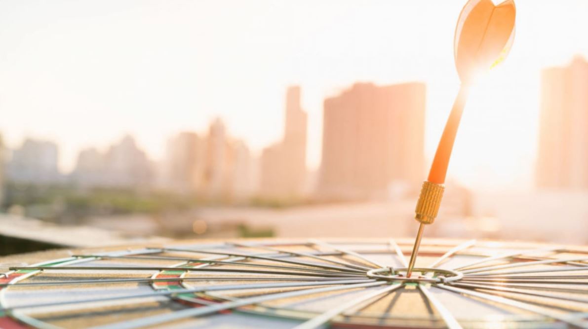 A dart hits the bullseye on a dartboard with a city skyline in the blurred background under a sunny sky.