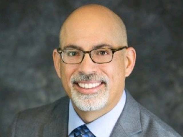 A person with glasses, wearing a gray suit and blue tie, smiles against a dark background.