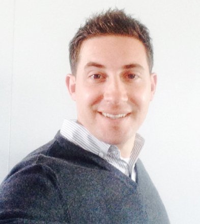 A smiling person wearing a dark sweater and collared shirt poses against a plain white background.