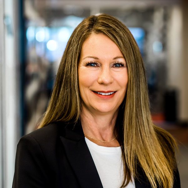 A woman with long hair, wearing a black blazer and white shirt, smiles in a well-lit indoor office setting.