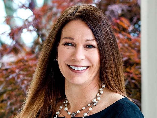 Smiling woman with long brown hair, wearing a pearl necklace and black top, standing near reddish foliage.